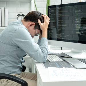 Male programmer feeling headache and stress during checking prog Male programmer feeling headache and stress during checking program code and debugging on multiple screens while he is programing to developing app and website of corporate at modern software office.